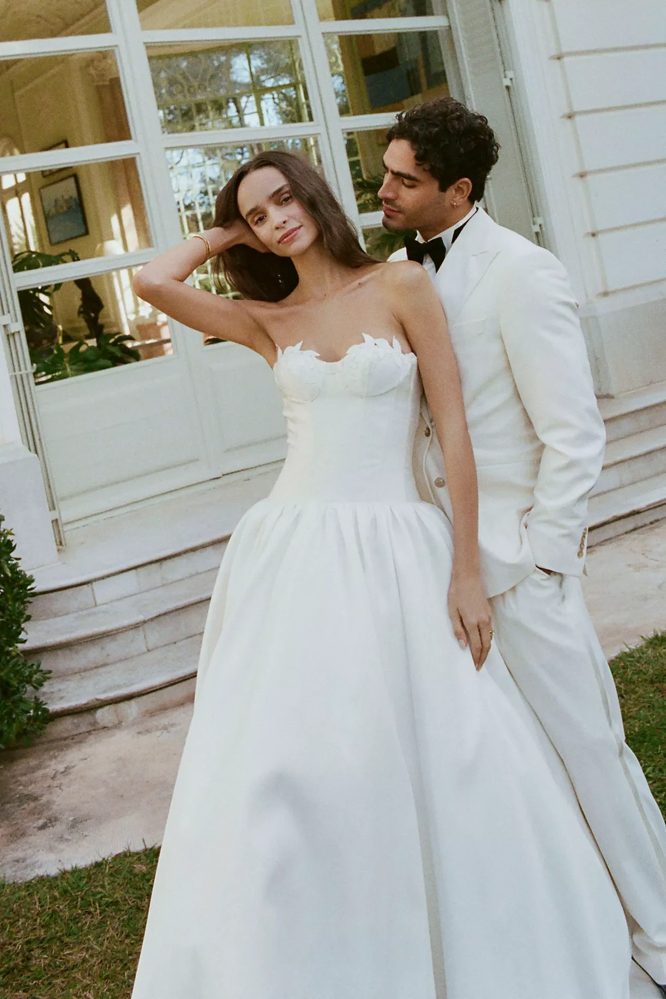 A woman in a white strapless wedding gown standing outside beside a man in a white tuxedo. The woman has dark hair and is touching her hair with her left hand, and the man has dark curly hair and is looking at her. They are in front of a house with large glass windows.