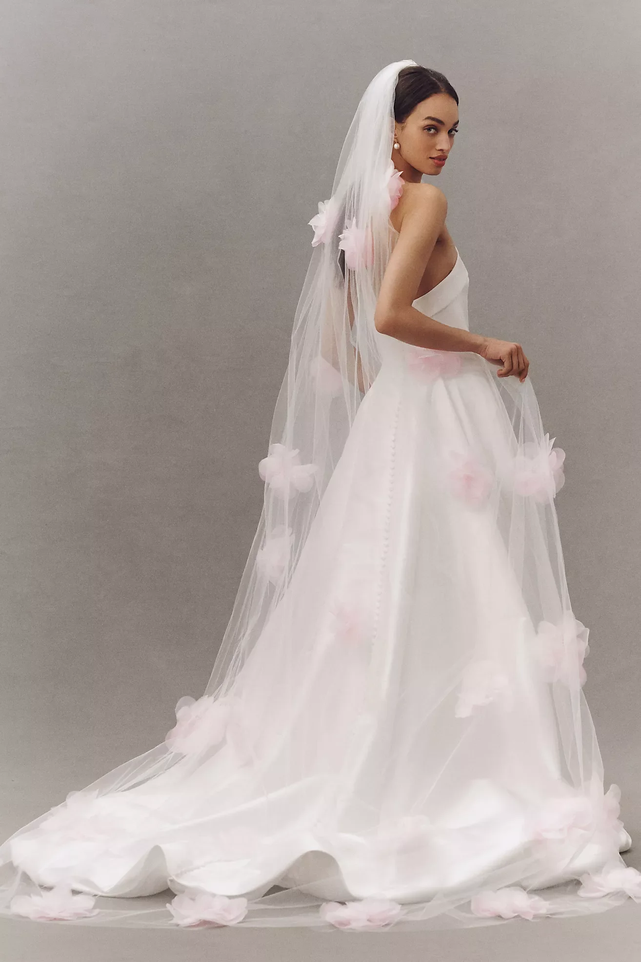 A woman in a wedding dress with a long, flowing train and a veil stands against a plain gray background.