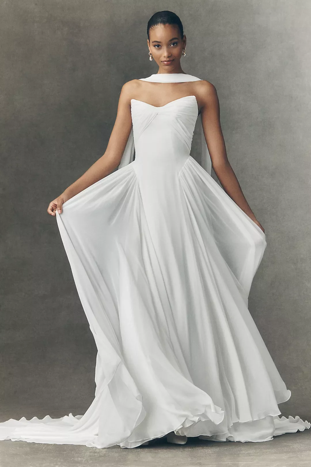 A woman in a white wedding dress with a fitted bodice and flowing skirt, standing against a plain gray background, slightly lifting the skirt with her right hand.
