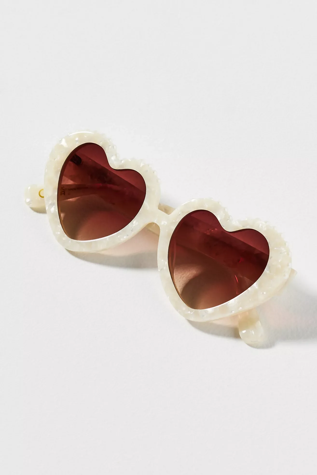 A pair of white shell-shaped sunglasses with red-tinted heart-shaped lenses on a white surface.