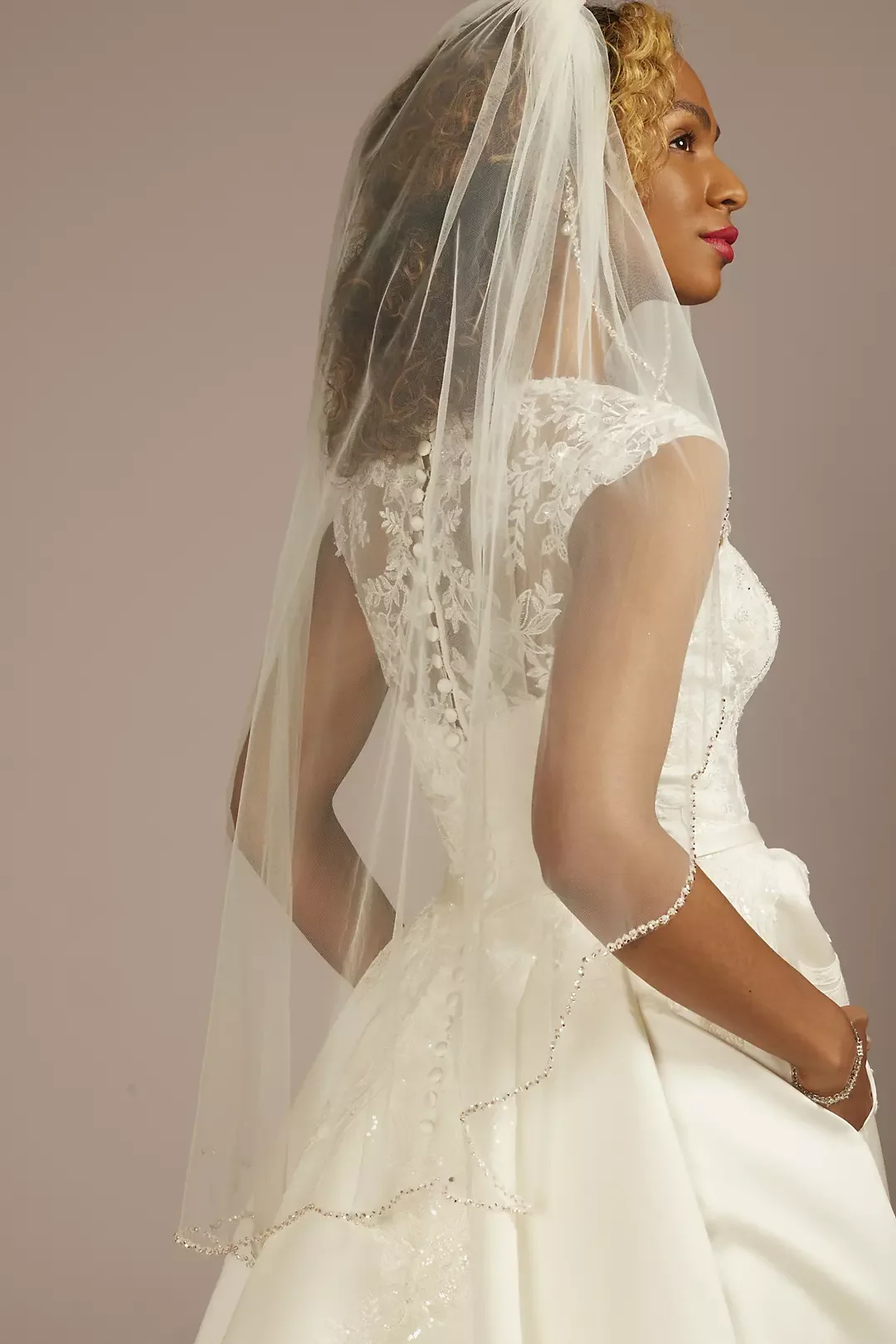 A woman in a wedding dress with a veil over her face, facing sideways with a neutral background.