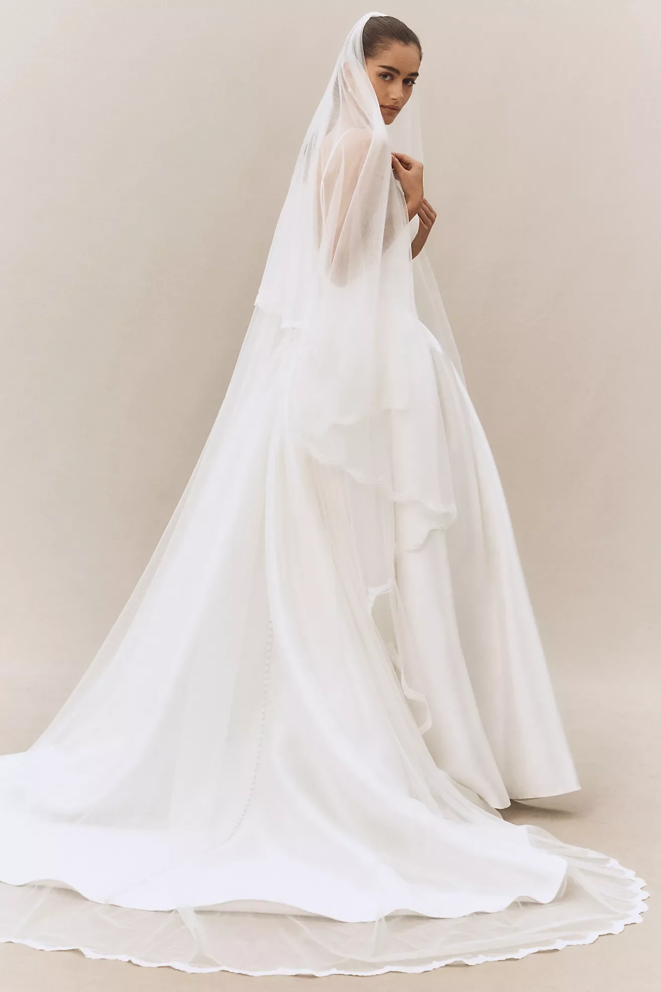 Woman wearing a long, flowing white dress with a sheer veil, standing against a plain light-colored background.