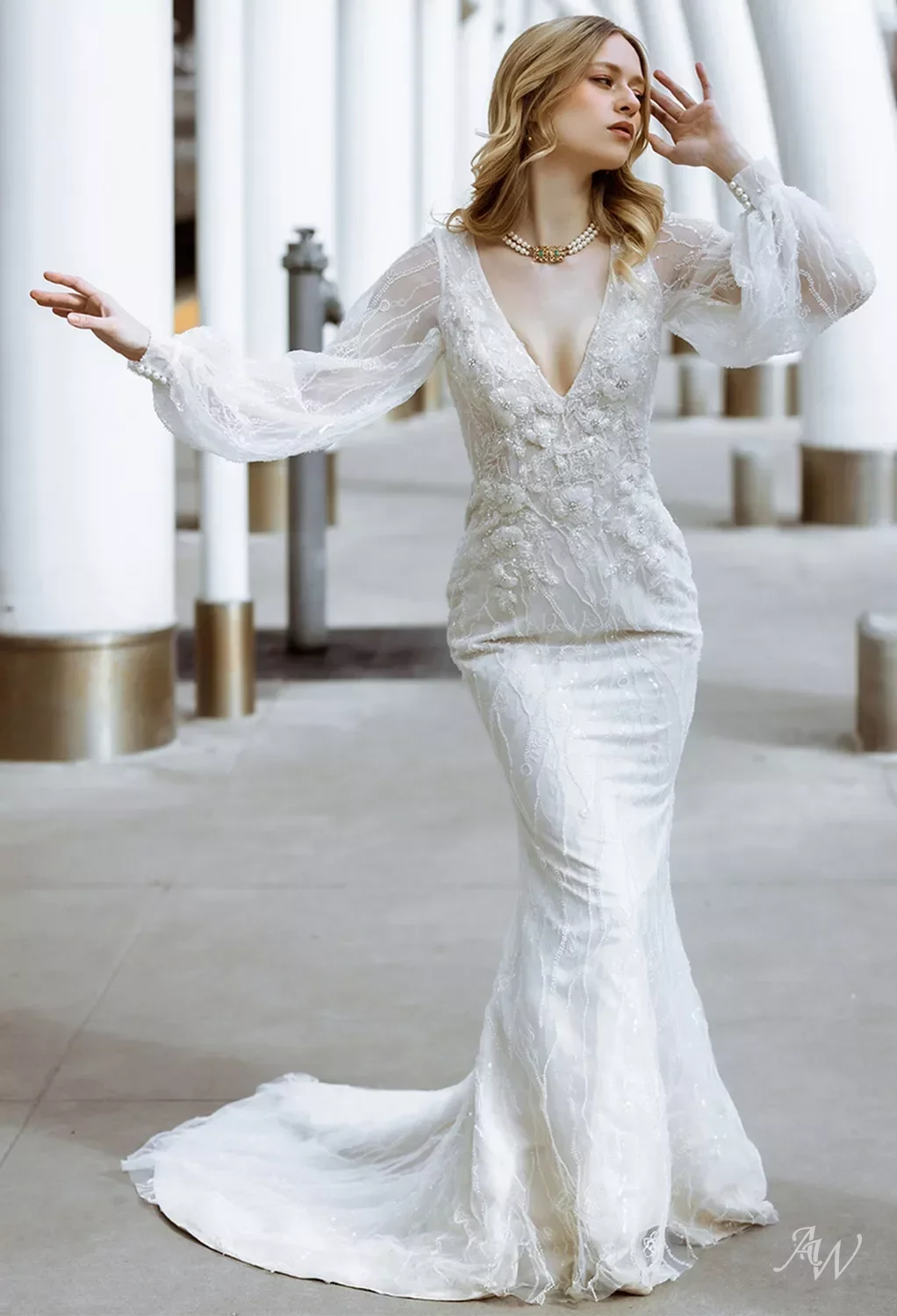 A woman in a white lace wedding dress with long puffy sleeves, standing outdoors in front of white columns and gold accents, with her hand raised near her face.