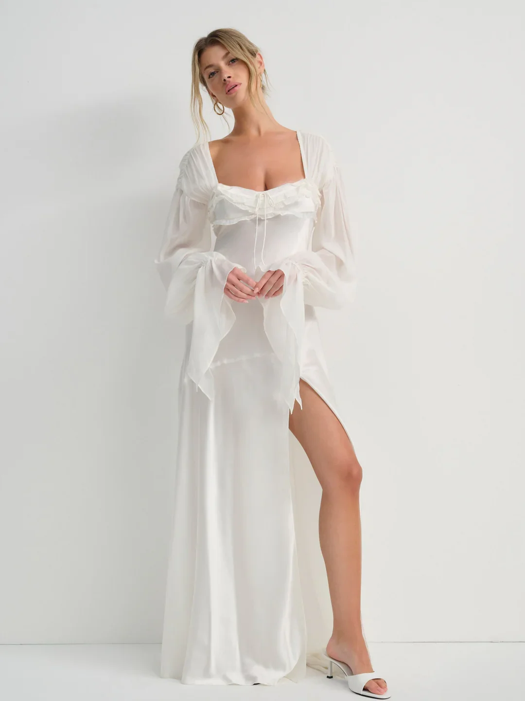 A woman in a long, elegant white gown with a thigh-high slit, standing against a plain white wall, wearing white high heels, with her hands in front of her.