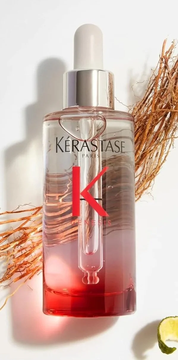 A bottle of Kérastase Paris hair treatment with a dropper cap, placed on a white surface with some dried plants and a slice of lime nearby.