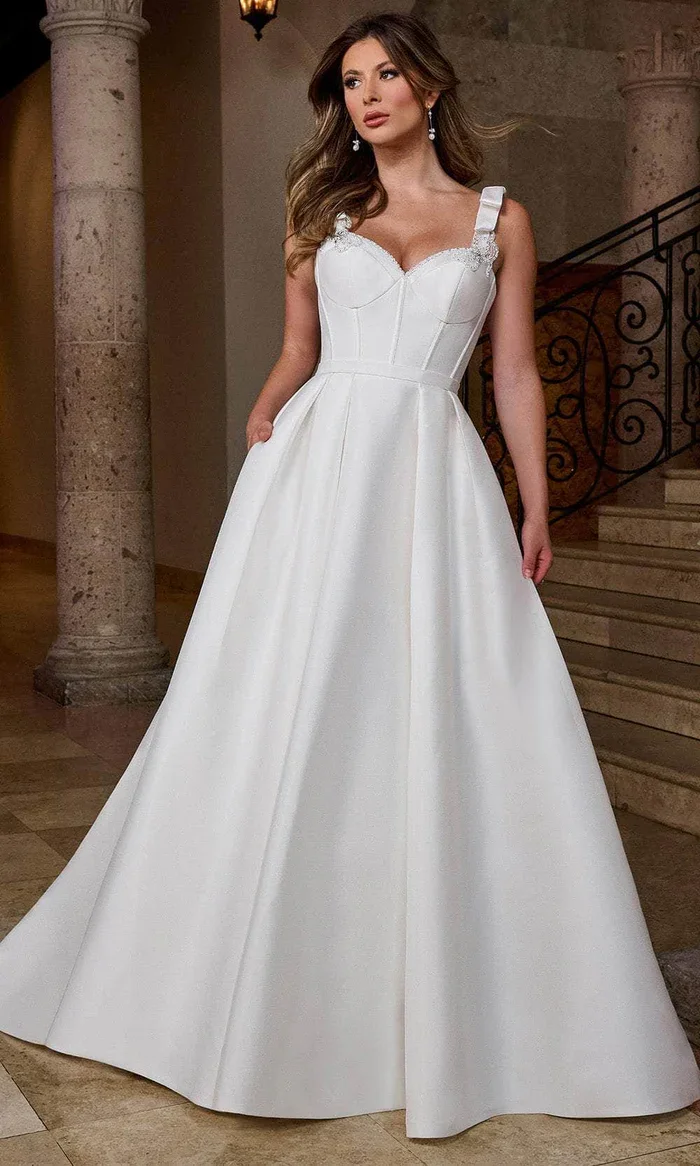 A woman in a white wedding dress with a fitted bodice, wide straps, and a full skirt stands indoors near stone columns and a staircase.