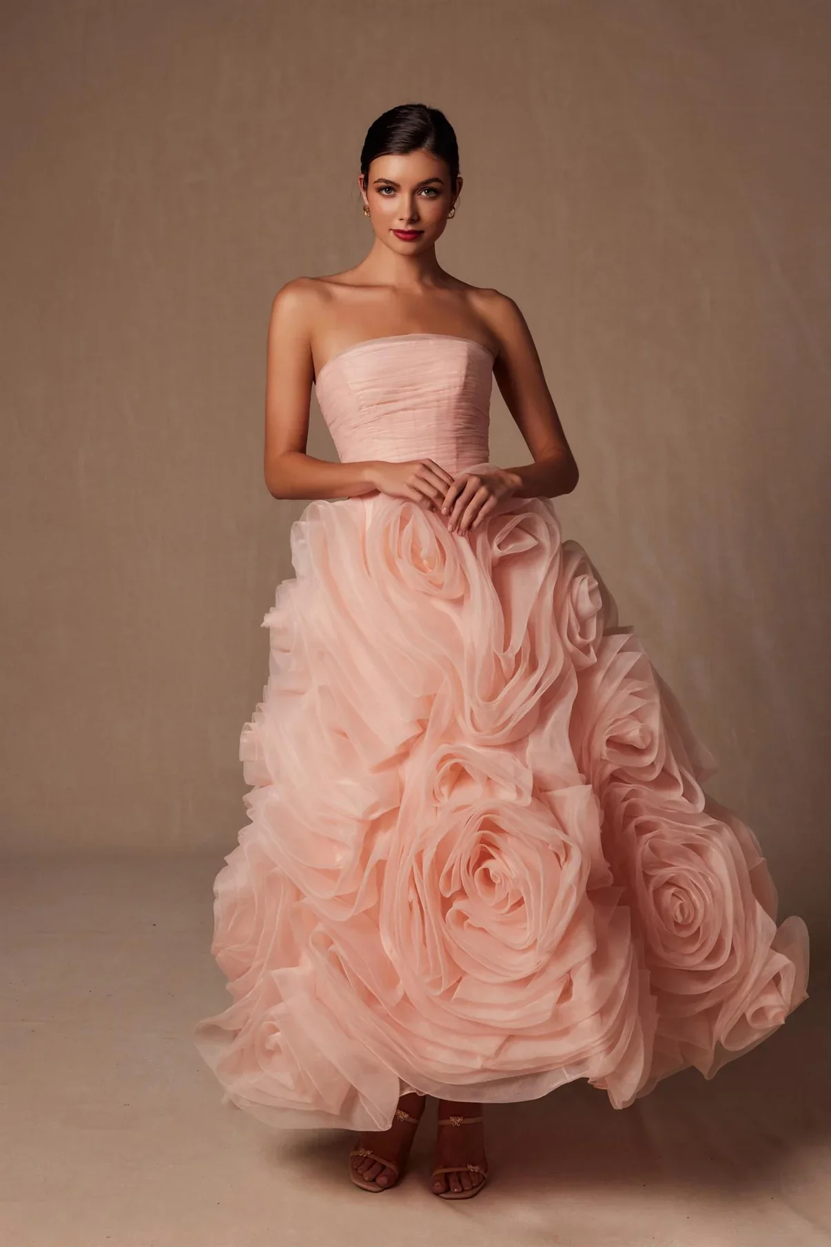 A young woman in an elegant, strapless peach-colored gown with a fitted bodice and a voluminous, ruffled skirt featuring rose-like fabric details, standing against a neutral background.