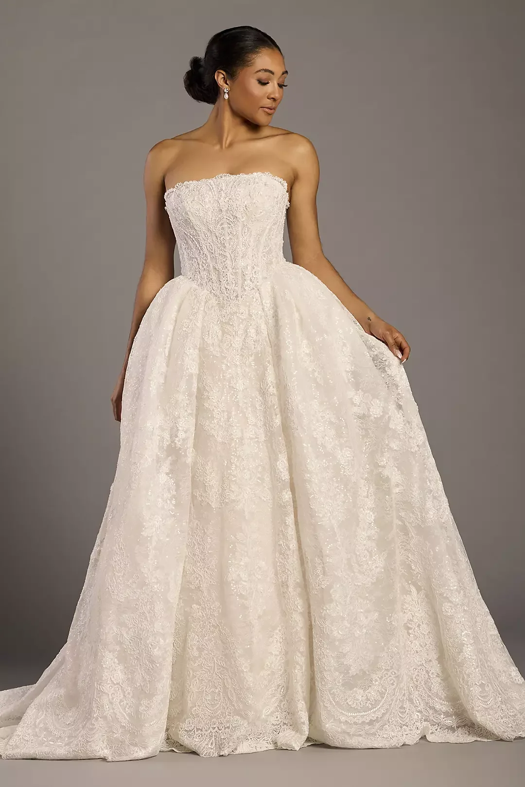 Woman wearing a strapless white lace wedding dress with a full skirt and fitted bodice, holding fabric and standing against a gray background.