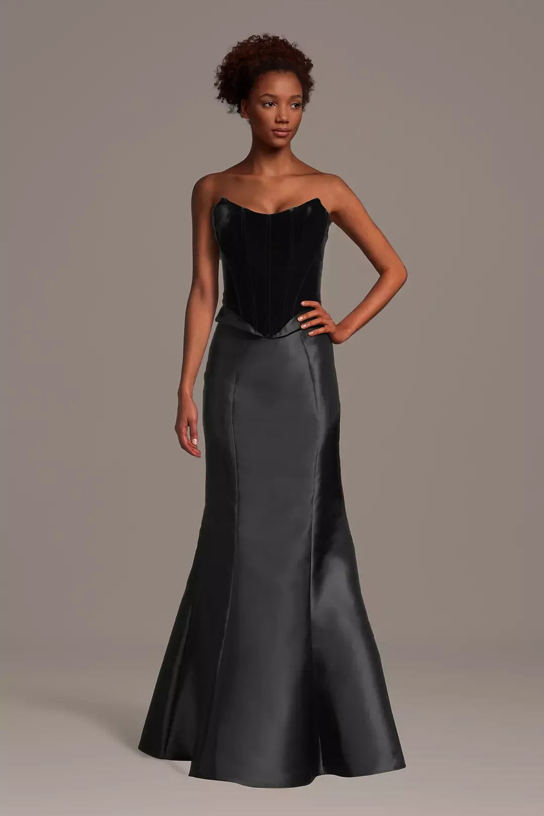 A woman with brown, curly hair wearing a strapless black velvet corset and a long, fitted black satin skirt that flares at the bottom.