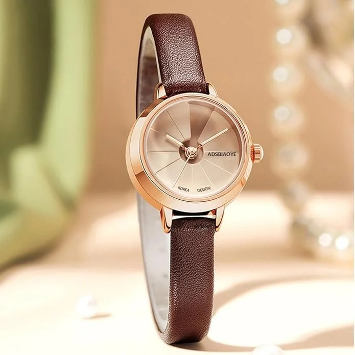 A round wristwatch with a gold-tone case, a metallic beige dial, and a brown leather strap on a light surface. The watch has minimal markings and three hands, with the brand name ADASBIAOYE on the dial.