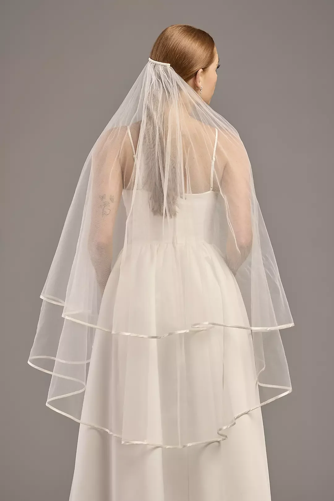 A woman in a white wedding dress with a layered skirt and a sheer veil standing with her back to the camera against a gray background.