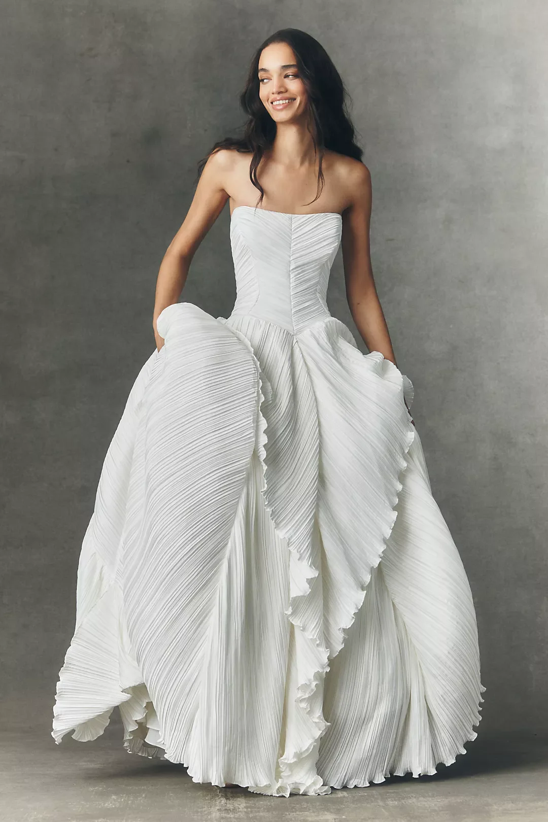 A woman in a white strapless gown with pleated fabric and ruffled details standing against a gray background.