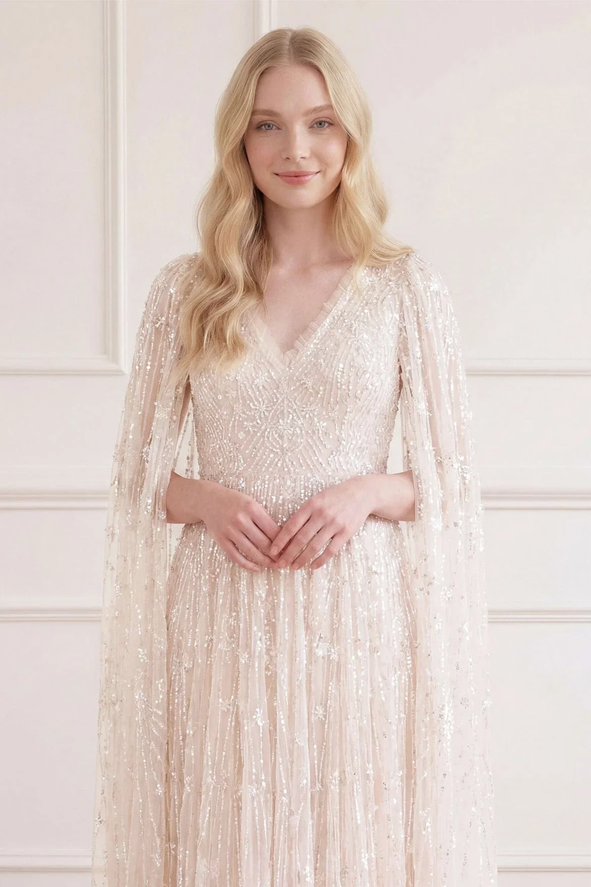 A woman with long blonde hair wearing an elegant, sparkling, beige evening gown with a V-neckline and sheer, embellished cape sleeves, standing against a white wall with decorative paneling.