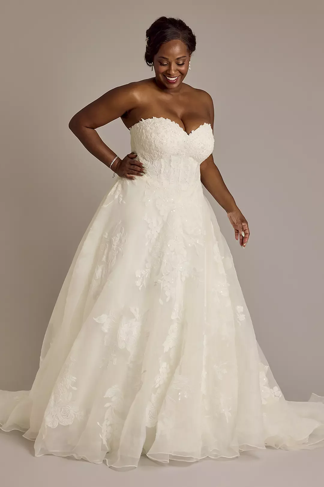 A woman wearing a strapless white wedding gown with lace details, smiling and looking down.
