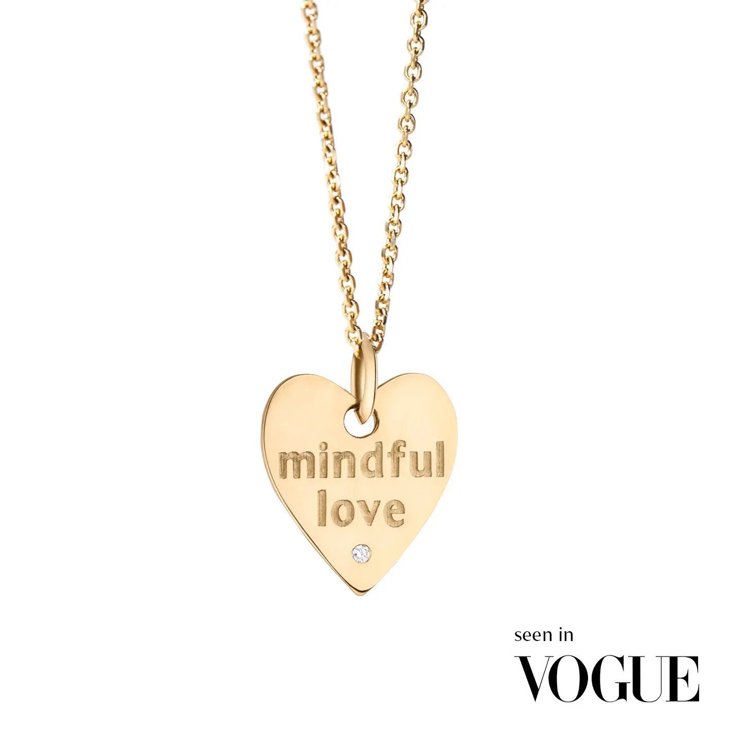Gold heart-shaped necklace with engraved words 'mindful love' and a small gemstone at the bottom, against a white background, Vogue logo in the corner.