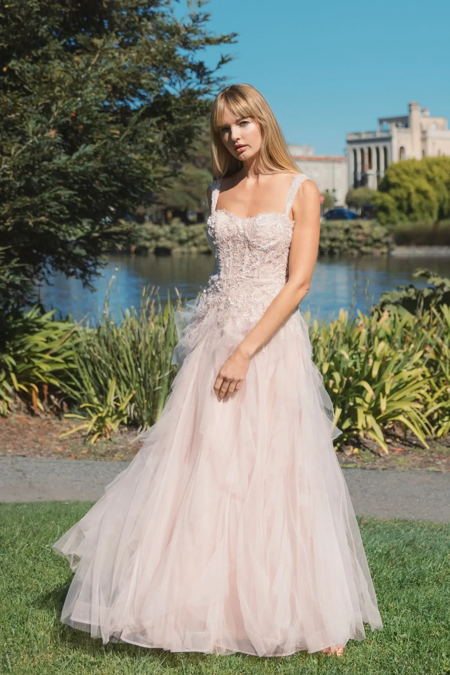 A young woman in a light pink wedding dress stands outdoors near a pond with trees and buildings in the background.