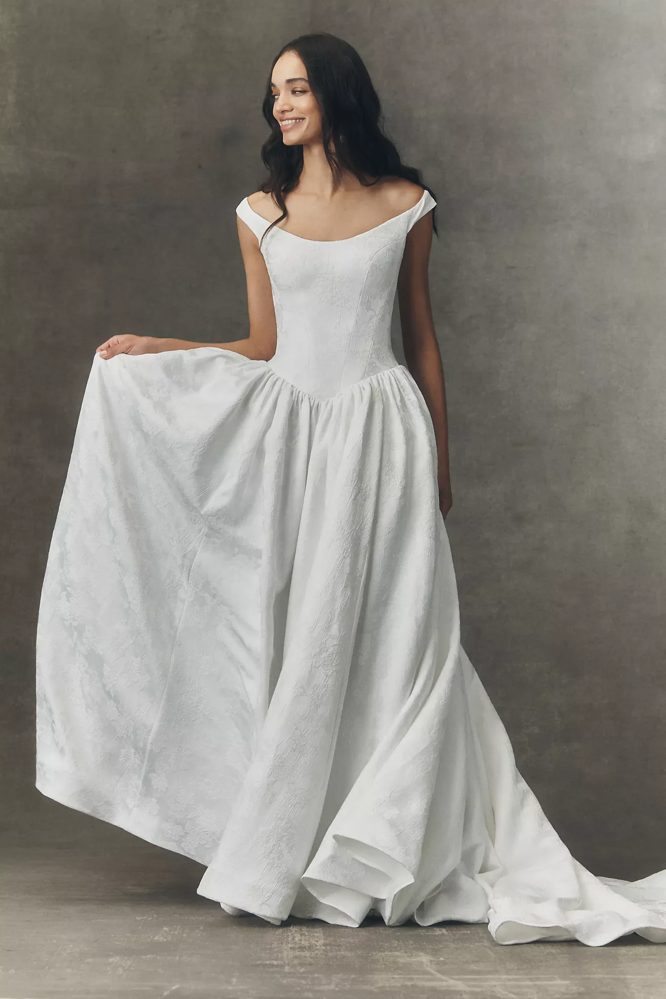A woman in a white wedding gown, smiling, holding the side of her dress, standing against a plain gray background.