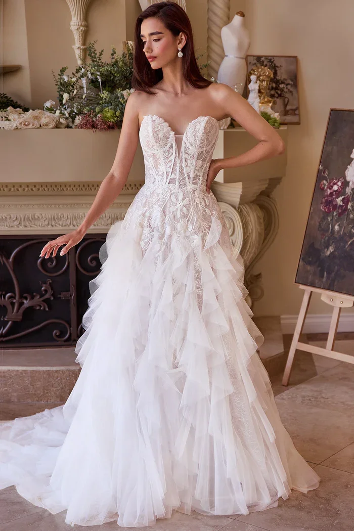 A woman modeling a strapless white wedding gown with a lace bodice and layered tulle skirt in an elegant room with floral decor and artwork.
