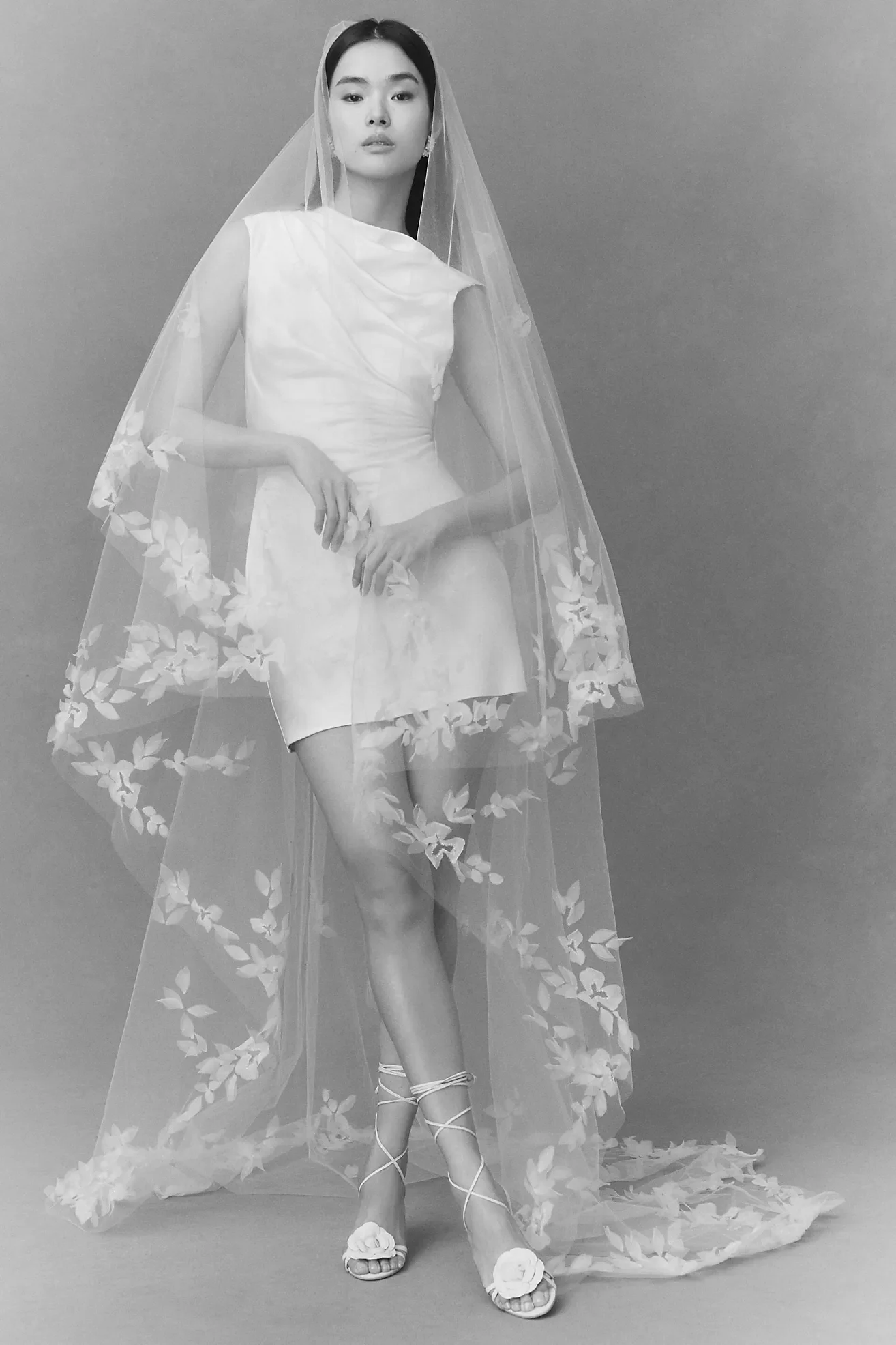 Black and white photo of a woman in a flowing dress with a sheer floral cape, wearing high-heeled sandals with floral accents, standing against a plain gray background.