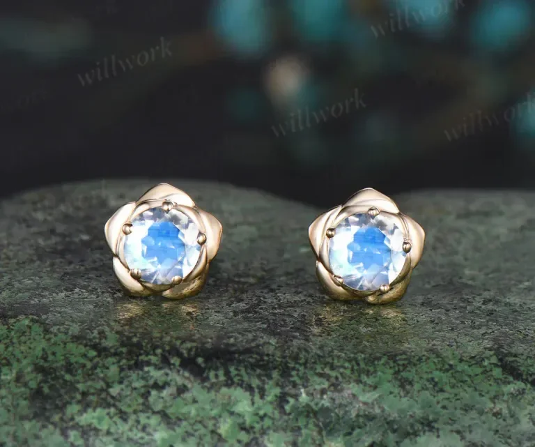 A pair of gold flower-shaped earrings with light blue gemstones, placed on a textured dark green stone surface.