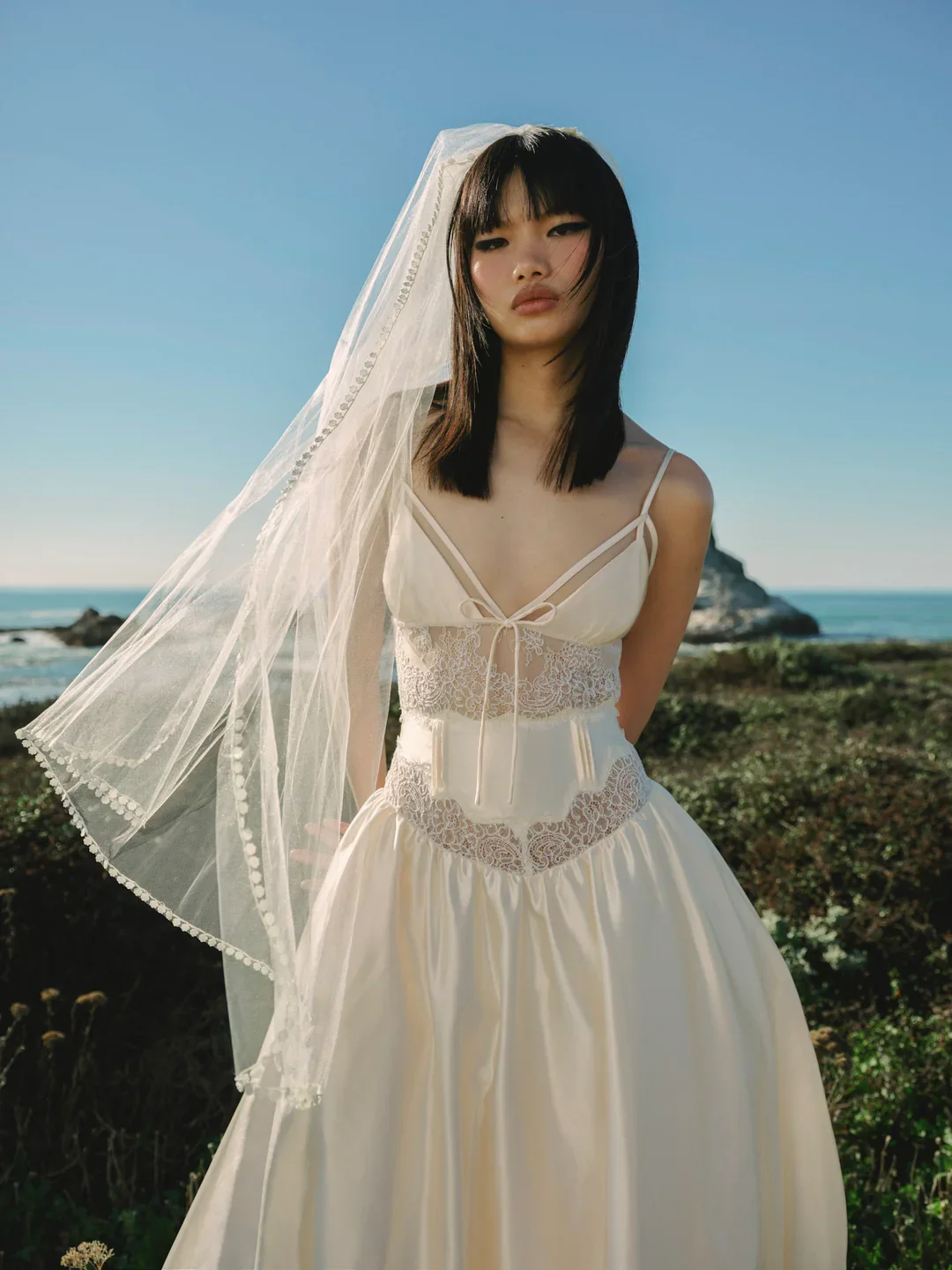 Young woman in a cream-colored dress and veil standing outdoors near the ocean with rocky cliffs in the background.