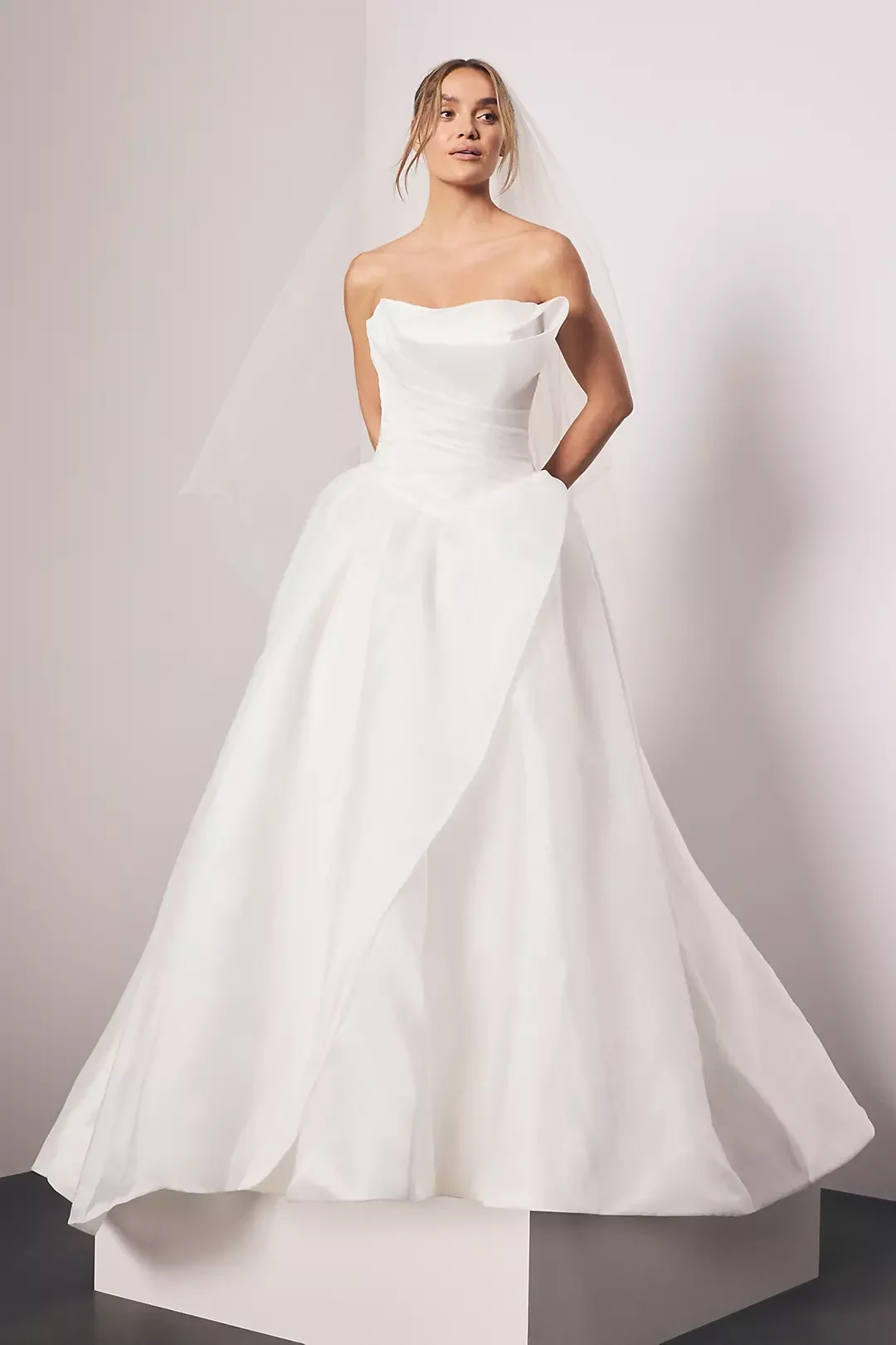 A woman in a strapless white wedding gown with a full skirt, standing against a plain background.