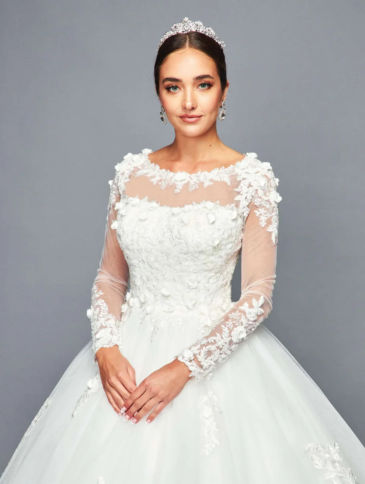 A woman in a white wedding gown with floral lace details, wearing a tiara and earrings, standing against a gray background.