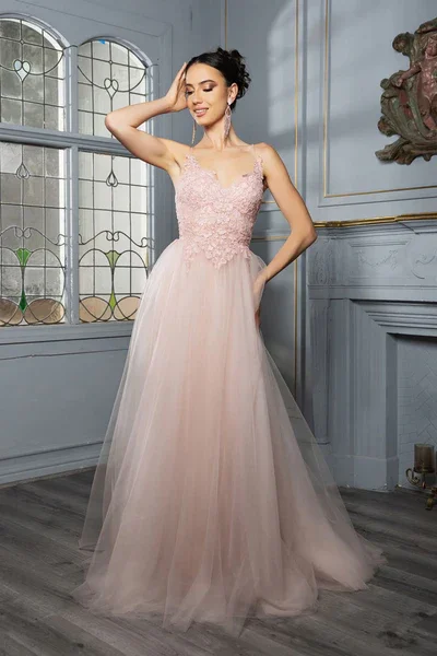 A woman in a pink, sleeveless tulle gown with lace detailing on the bodice, standing indoors near a window with stained glass design and decorative wall molding.