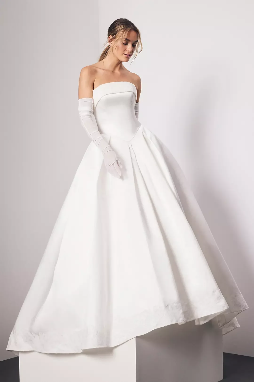 A woman wearing a strapless white wedding gown with elbow-length gloves, standing against a plain wall.