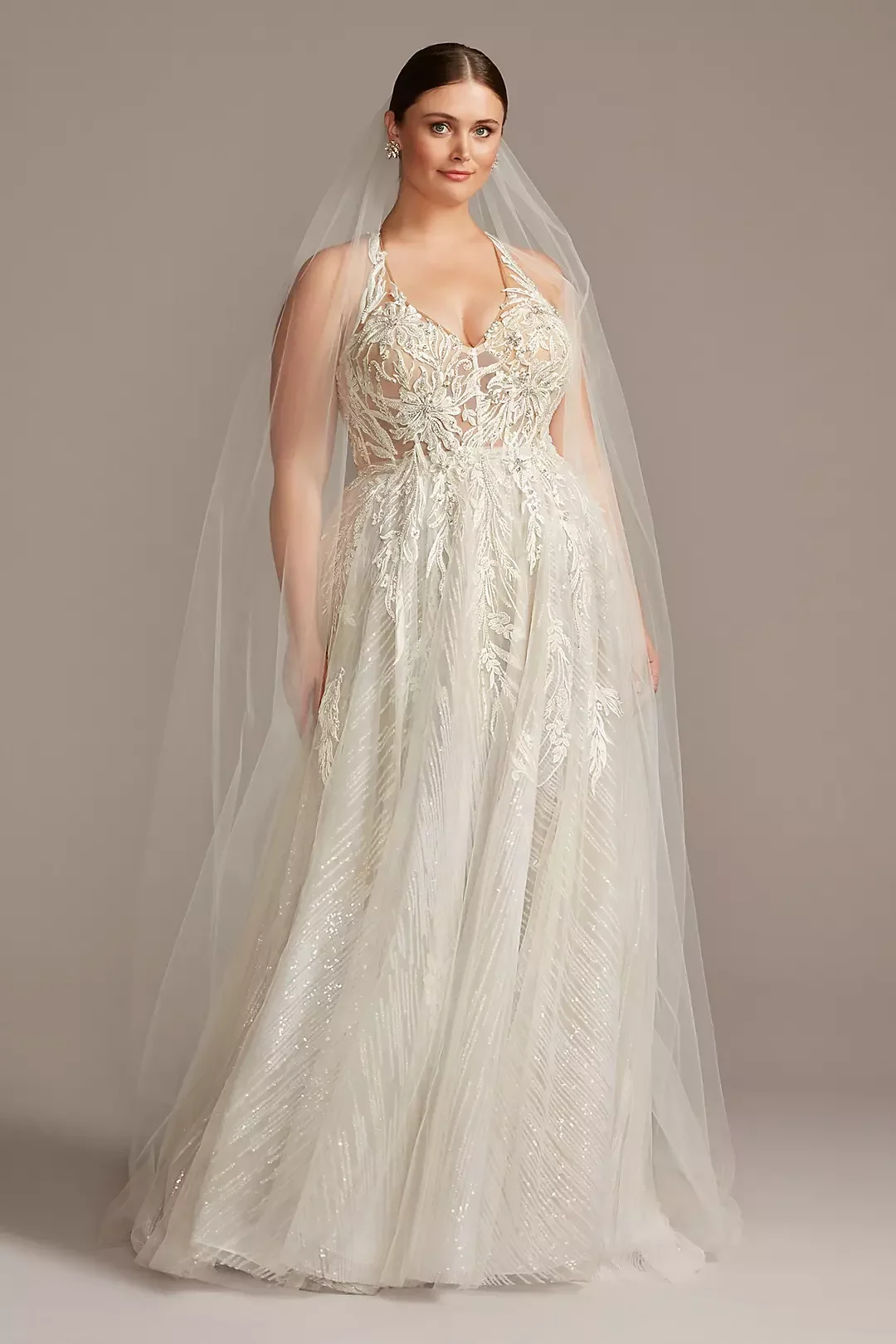 A woman in a detailed wedding dress with lace embroidery and a sheer overlay, standing against a plain background.