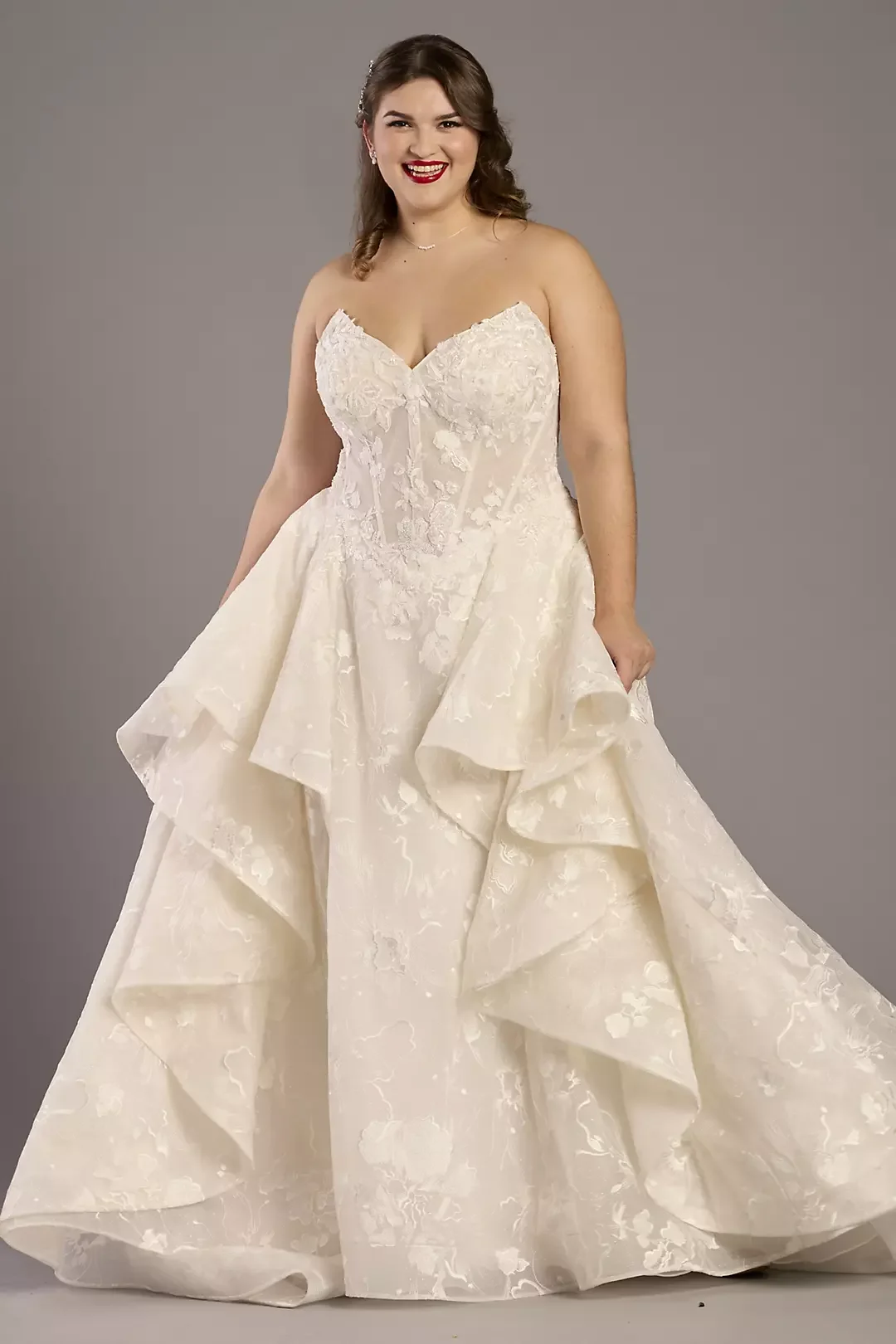 A woman in a white wedding gown with floral embroidery, smiling, holding the skirt with her right hand, standing against a gray background.