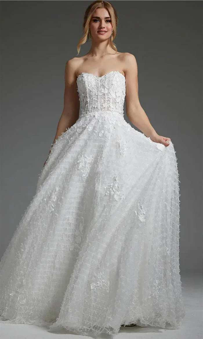 A woman in a strapless white wedding gown with textured lace details, holding the sides of her dress, against a gray background.
