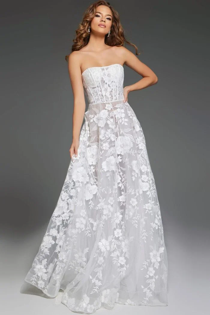 A woman in a strapless white lace wedding dress with floral patterns, standing against a gray background.