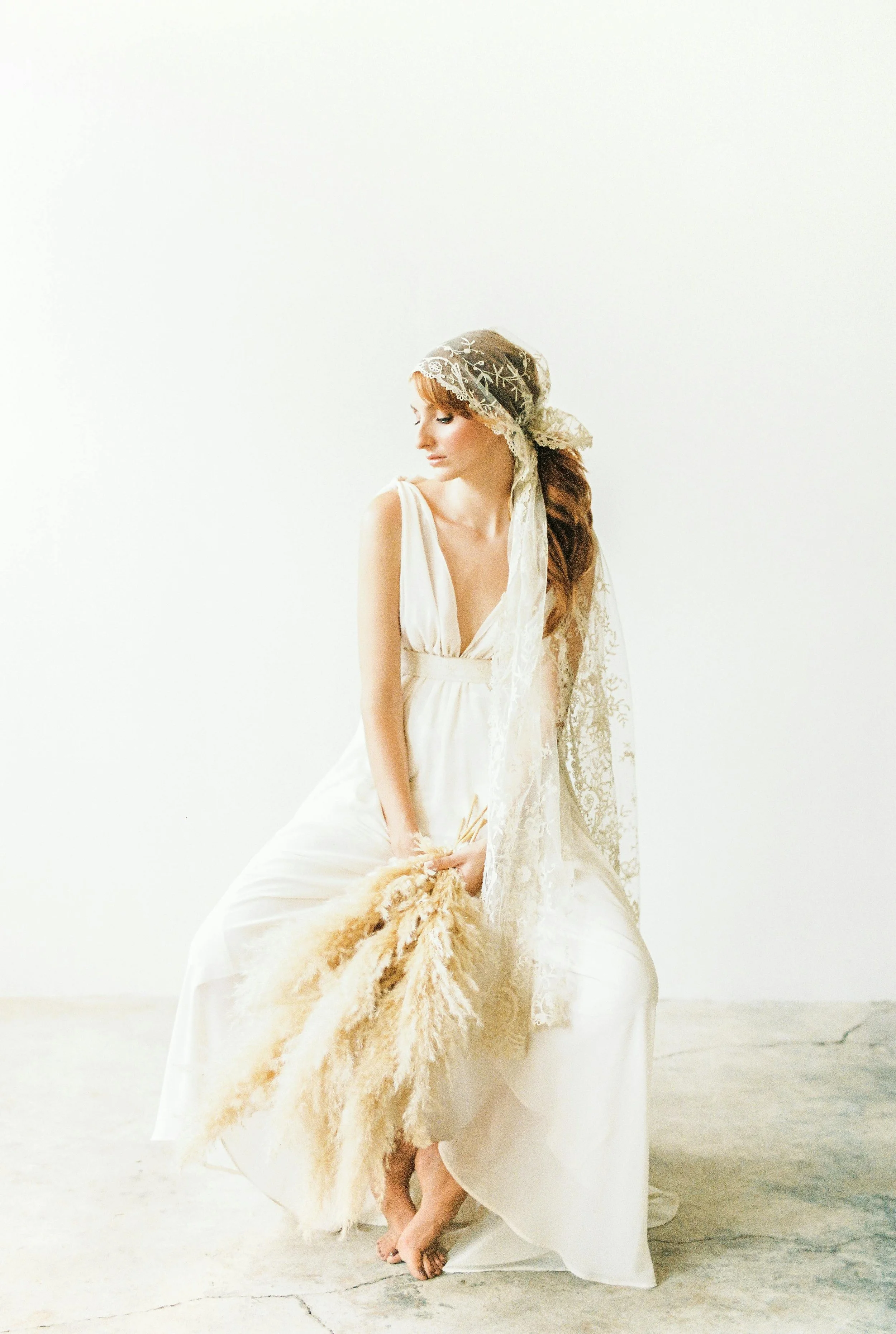 A woman in a white dress with a lace headscarf sitting on a concrete floor, holding a bunch of dried pampas grass.
