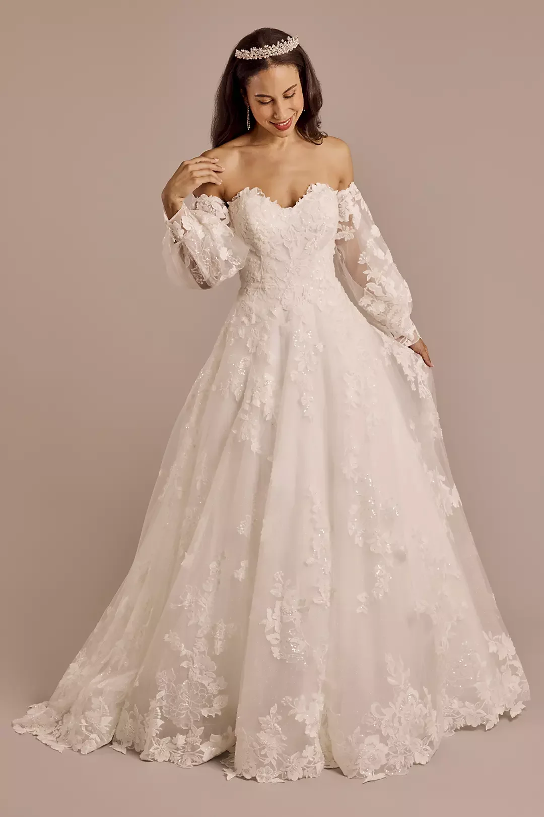 A woman in a white wedding dress with lace details, long sleeves, and a flowy skirt, wearing a tiara and earrings, smiling and holding her dress.