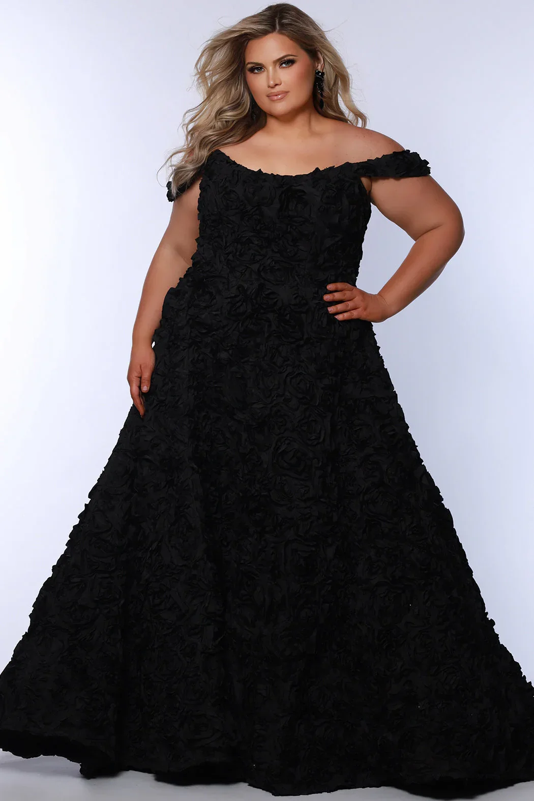 A woman wearing a black off-the-shoulder gown with floral textured fabric, standing against a plain light background.