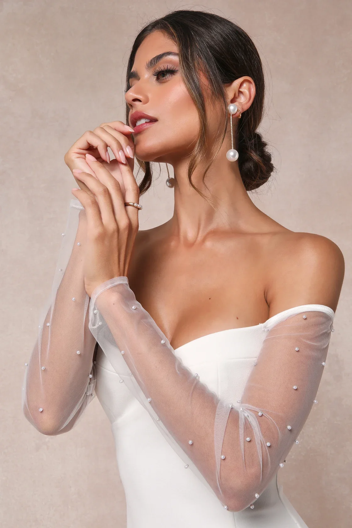 A woman with groomed hair and makeup wears a white off-shoulder dress with sheer, pearl-embellished sleeves, and large pearl earrings with dangling chains, posing with her hand near her face against a neutral background.