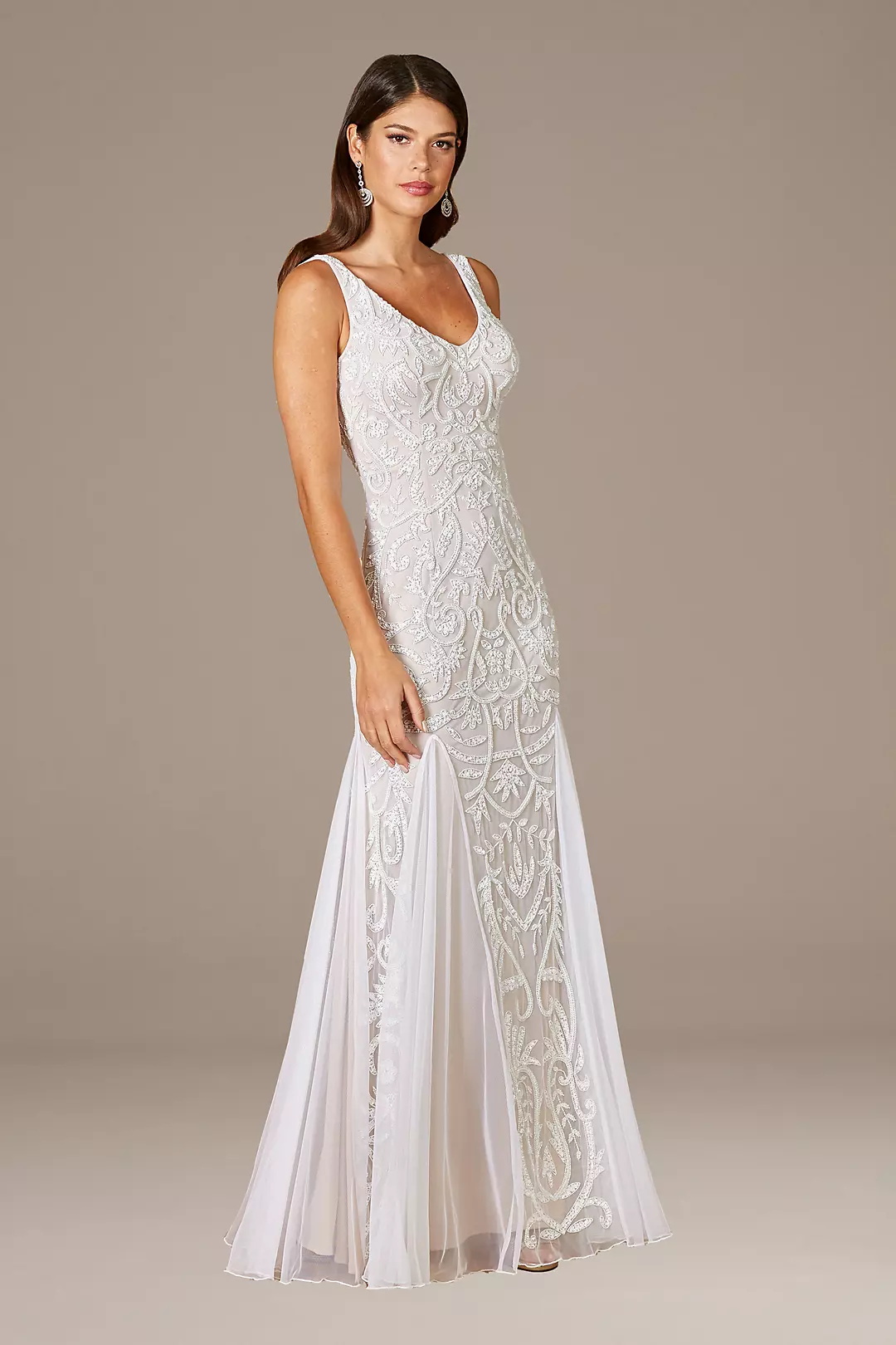 A woman in a white, sleeveless, lace wedding dress with intricate embroidery and a sheer overlay, standing against a neutral background.