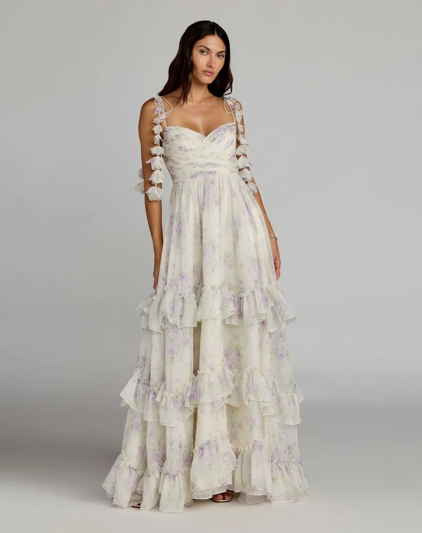A woman in a light-colored, floral, layered, floor-length dress with cold shoulder sleeves is standing against a neutral background.