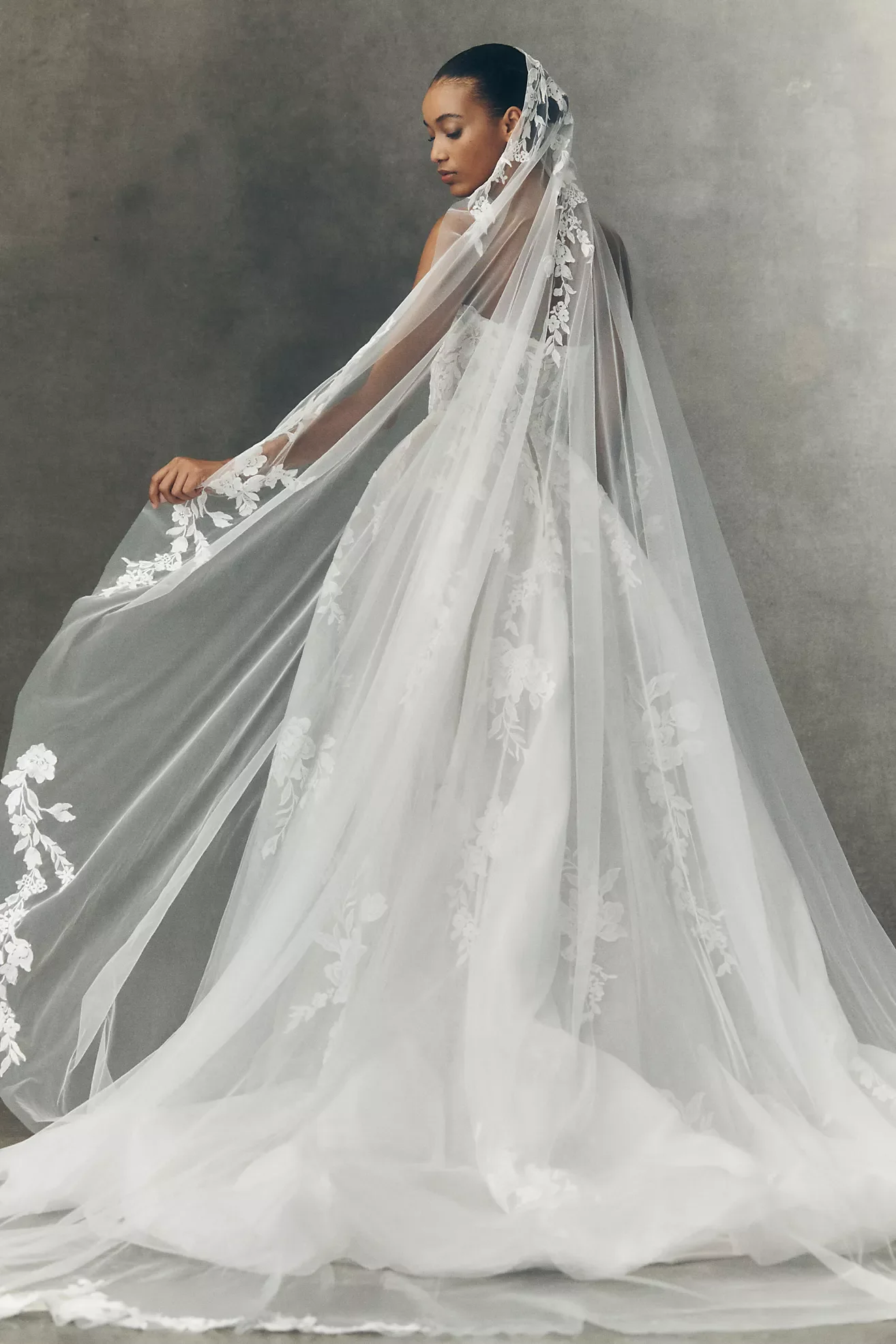 A woman in a wedding dress with a long, flowing train and lace details, standing against a plain grey background, holding a part of her veil.