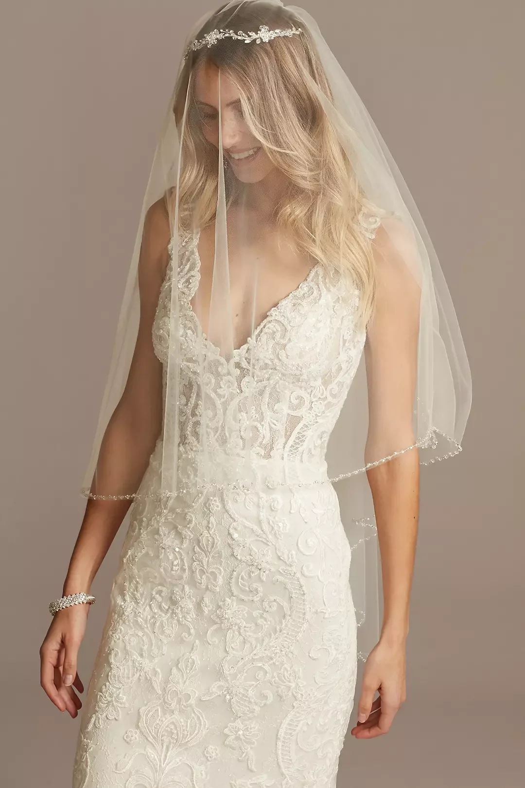 A bride wearing a white lace wedding dress with a veil and a tiara, smiling with her head tilted down.