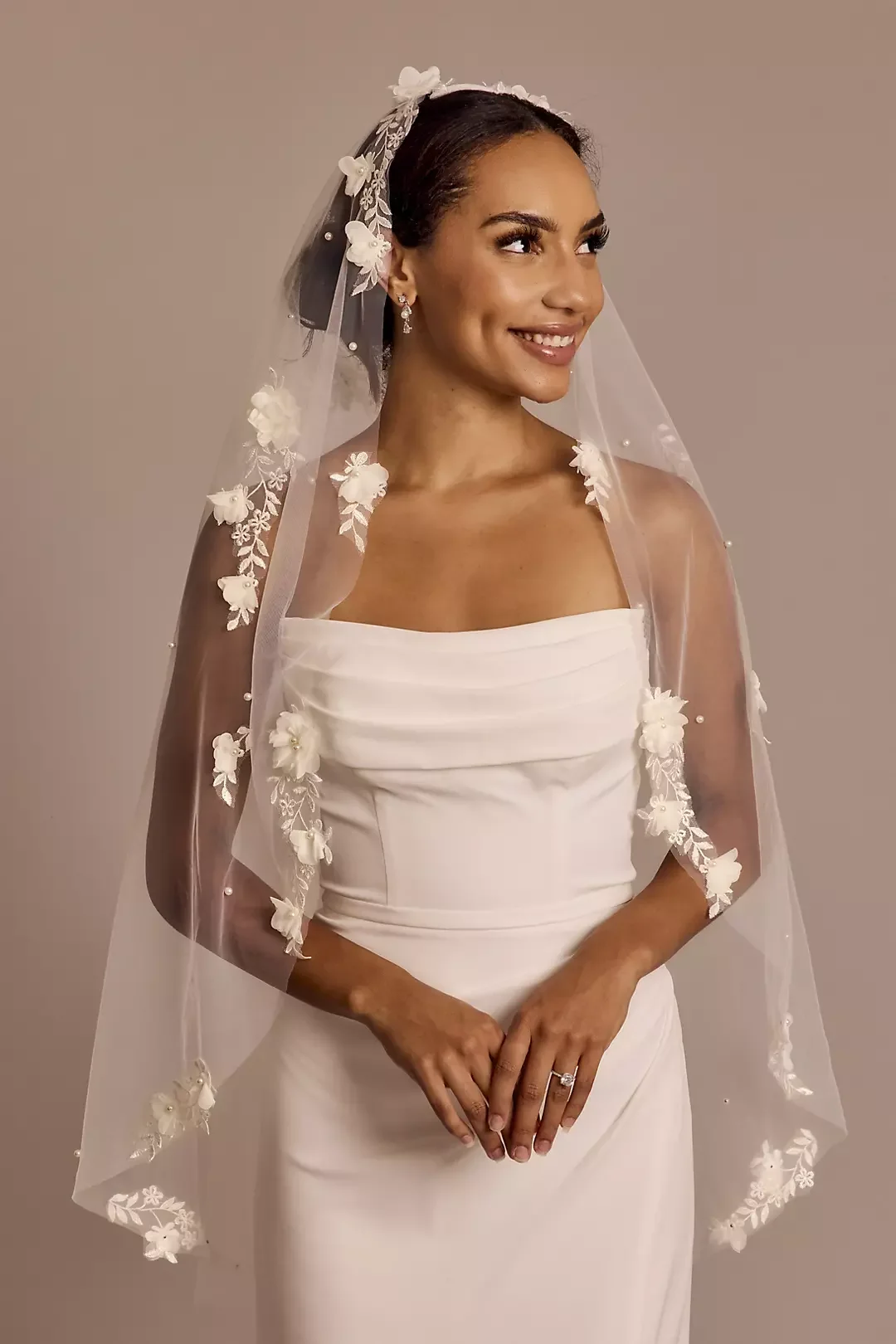 A woman in a white wedding dress with a veil decorated with flowers and lace, smiling and looking to her right.