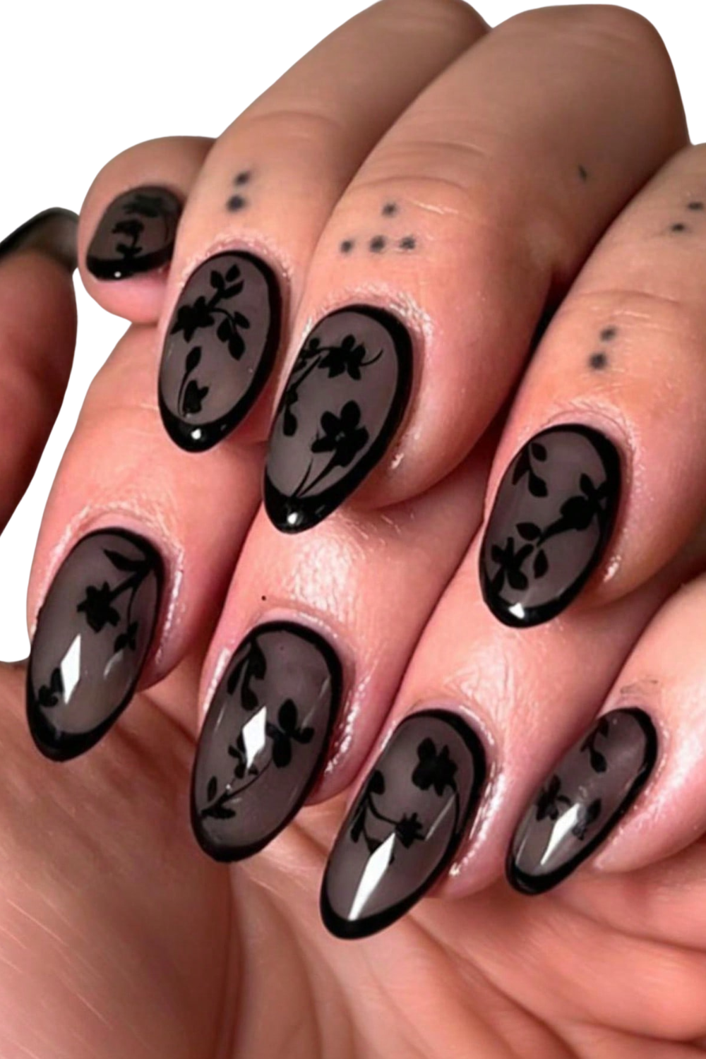 Close-up of a hand with black floral nail art on dark, glossy oval-shaped nails and small black dot tattoos on fingers.