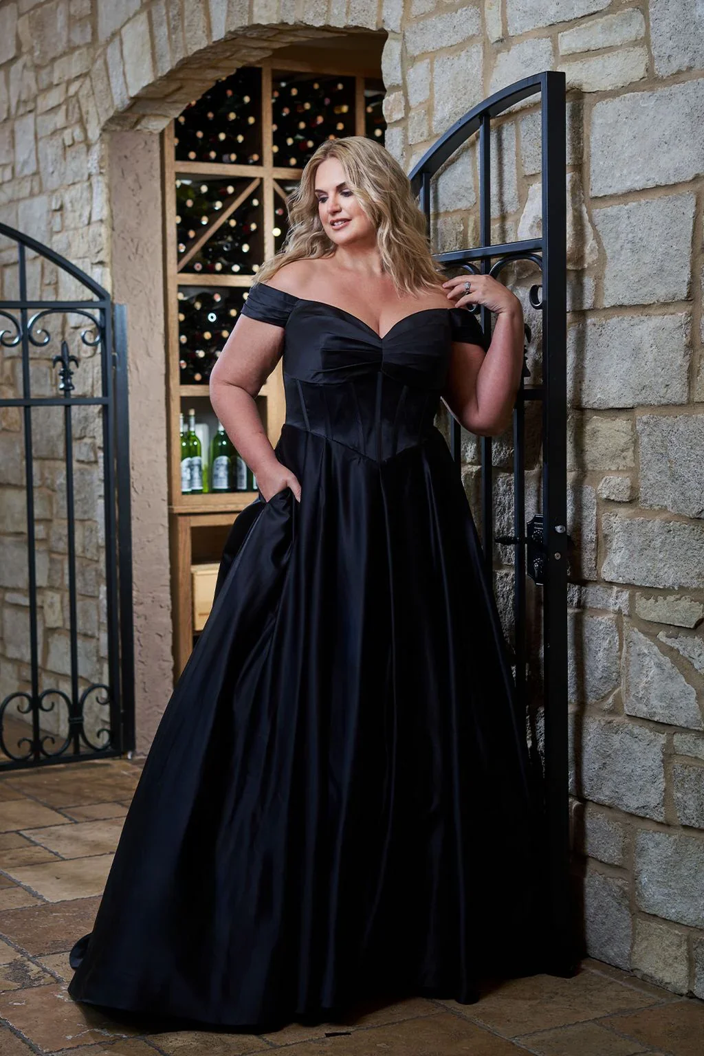 A woman in an off-shoulder black gown standing near a stone wall, with a wine rack and bottles in the background.