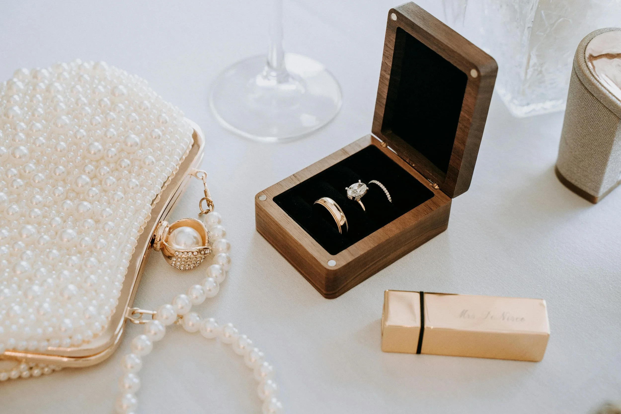 Wedding jewelry including pearl necklace, ring, and bracelet, a wooden box with rings and a diamond ring inside, a gold box, and a pearl-encrusted clutch on a white surface.