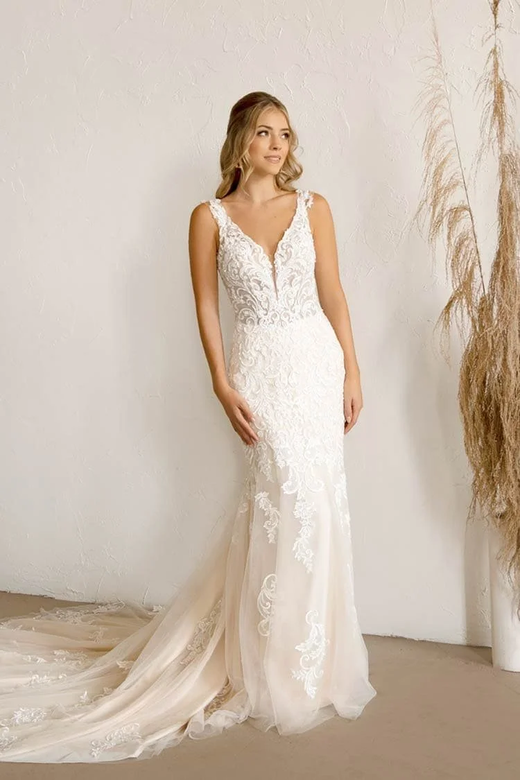 A woman wearing a white lace wedding dress with a train, standing indoors against a plain wall with dried plants on the side.