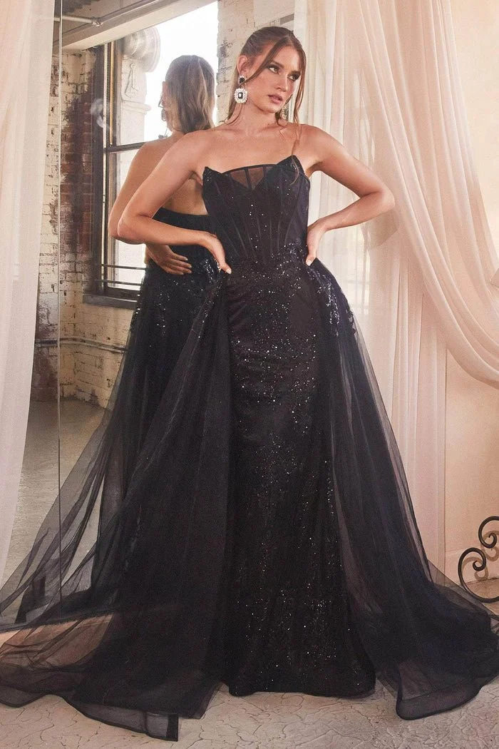 Woman in a black, strapless, sequined evening gown with a tulle overlay, standing in front of a mirror and curtains, posing with hands on hips.