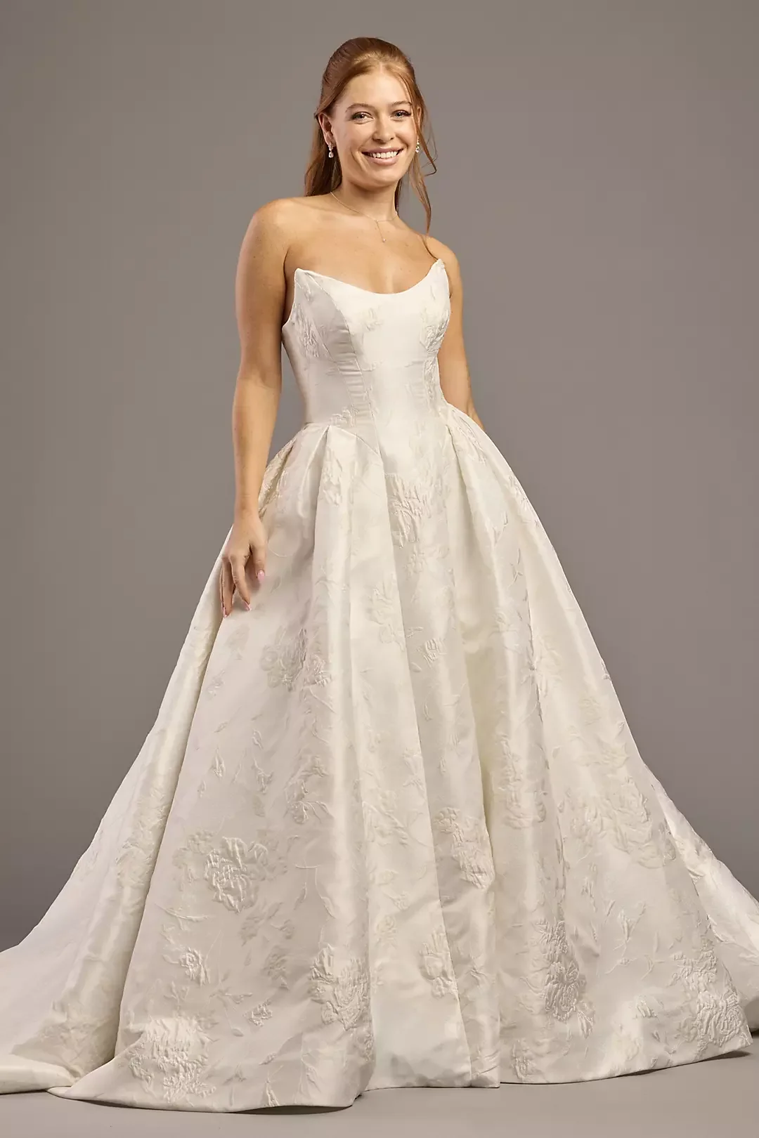 A woman with red hair smiling in a white strapless wedding dress with a full skirt and floral embroidery, against a plain gray background.