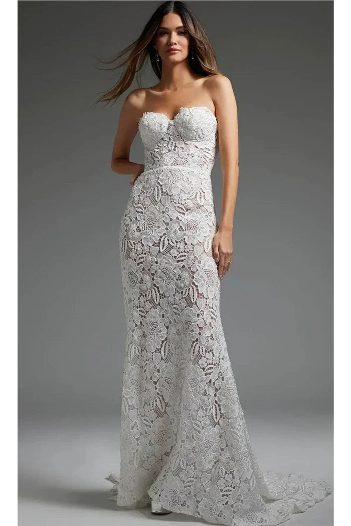 A woman in a white lace strapless dress. She has long brown hair and is standing against a gray background.