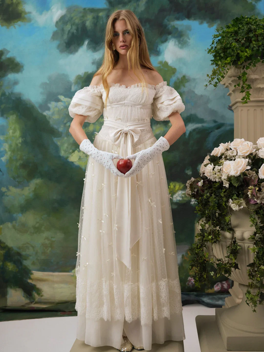 Young woman in a vintage white dress holding a red apple with both hands, standing in front of a painted landscape background, surrounded by flowers and greenery.